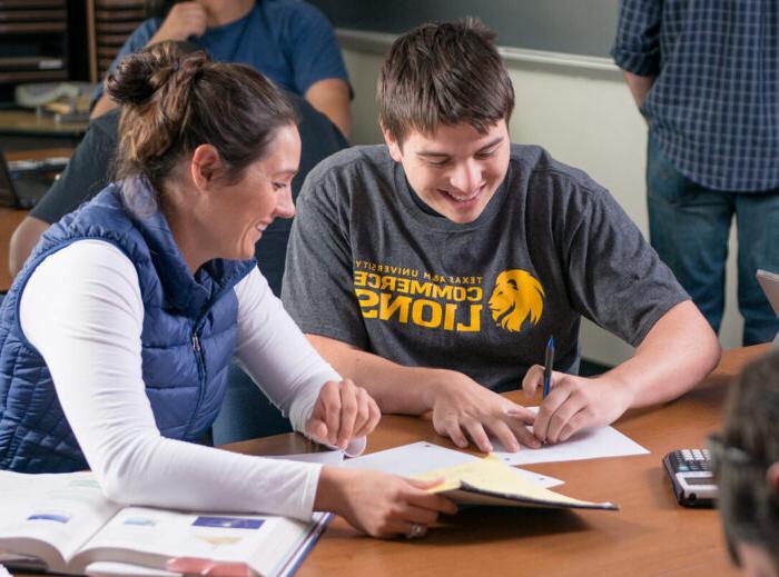 Two students work together at a table in a classroom. A student in the background is at the blackboard.